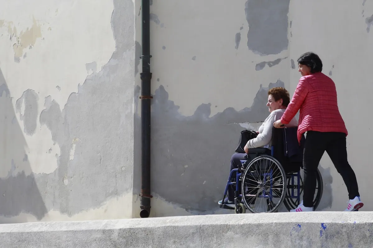 A person pushing a wheelchair user outdoors, representing physical accessibility and the curb cut effect.